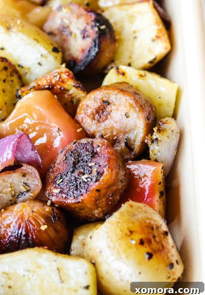 Close-up of golden brown roasted sausage, caramelized apple slices, and tender potatoes on a sheet pan, ready to serve.