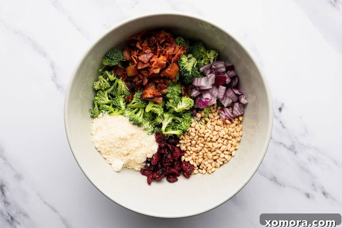 All the ingredients for the Easy Crunchy Parmesan Broccoli Salad laid out on a table, including chopped broccoli, red onion, cranberries, pine nuts, Parmesan, bacon, and dressing ingredients.