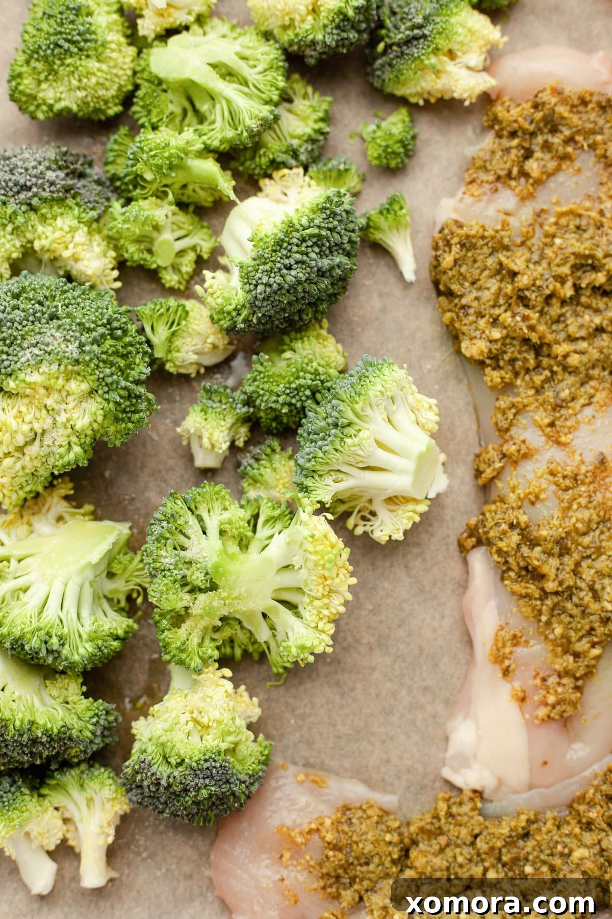 Pesto-coated chicken breasts and broccoli florets arranged on a baking sheet