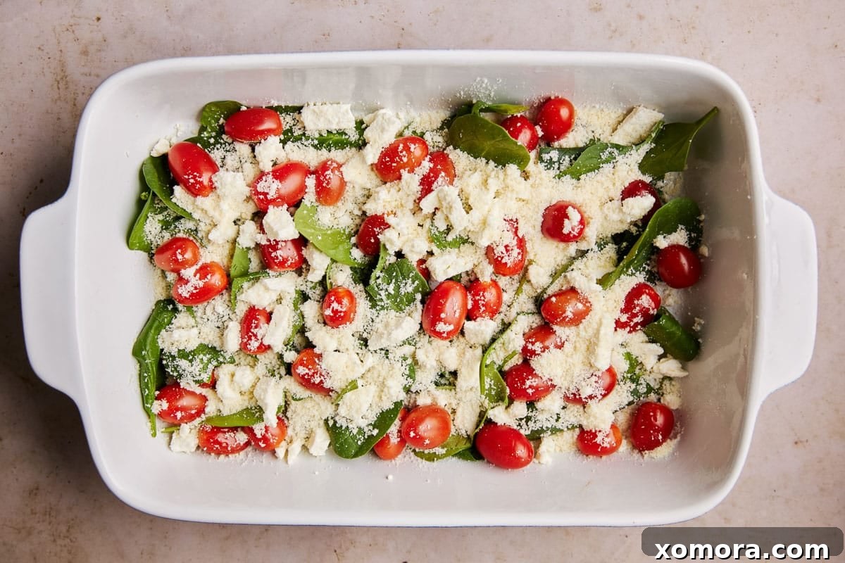 The baking dish with layers of spinach, grape tomatoes, crumbled feta, and grated Parmesan cheese, ready for the egg mixture to be poured over.