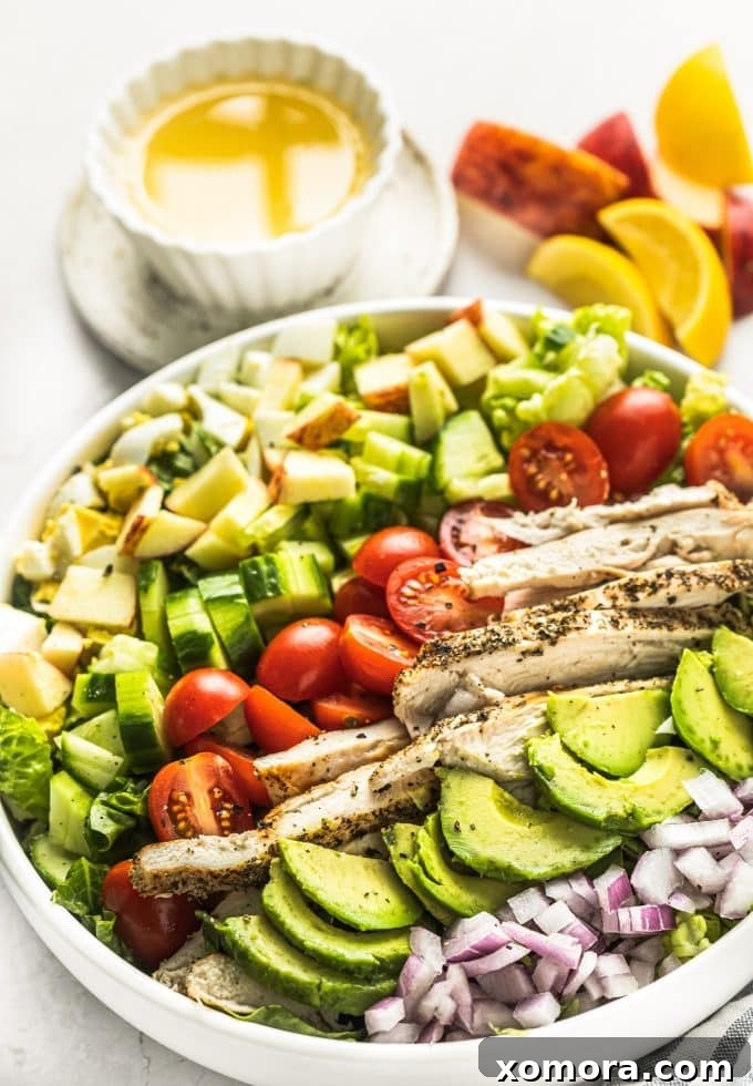 A vibrant Lemon Chicken Cobb Salad artfully arranged with crisp lettuce, sliced chicken, hard-boiled eggs, avocado, cucumber, red onion, and apple slices, drizzled with a golden lemon vinaigrette.